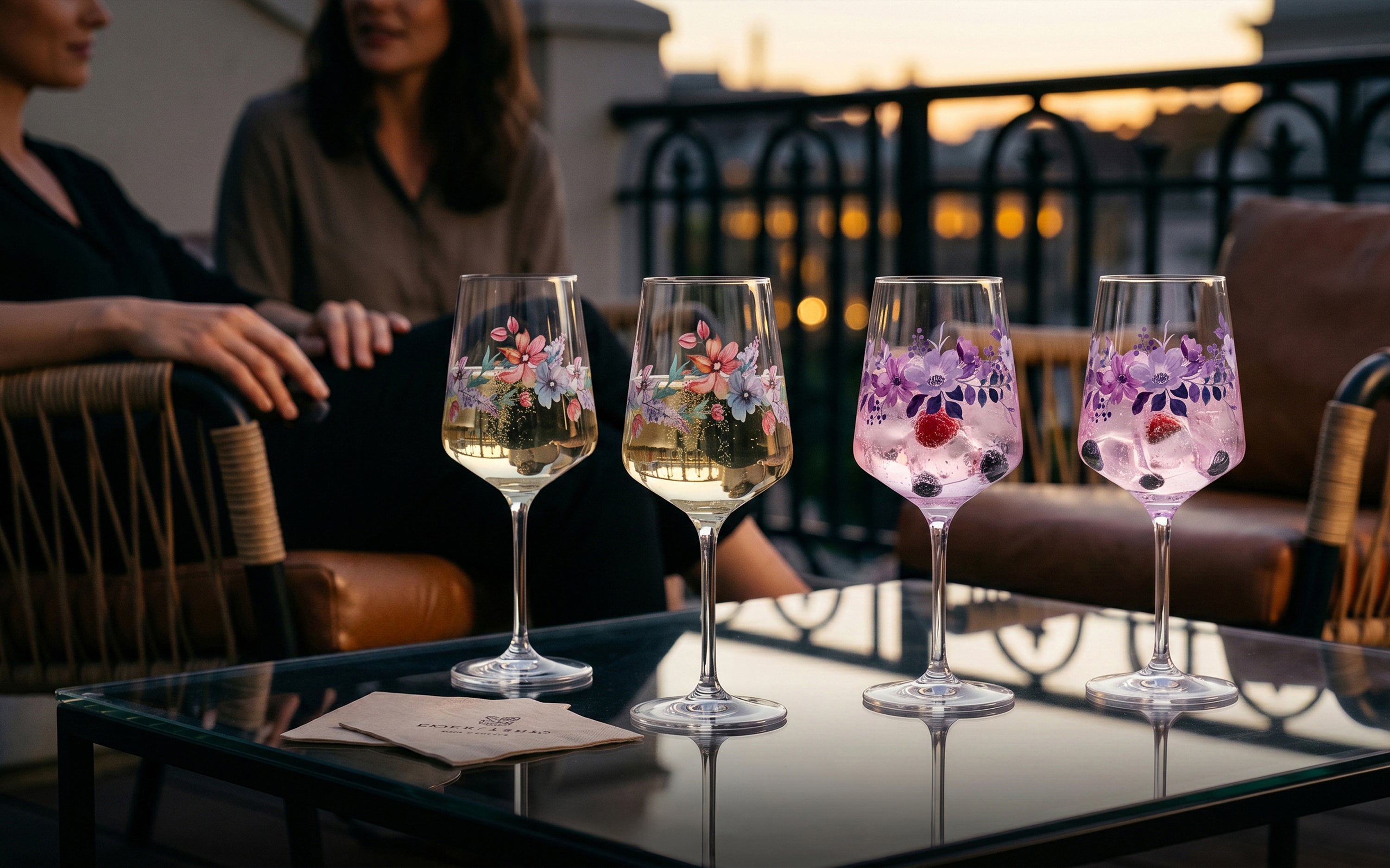 Vier ElbFuchs Aperitifgläser mit floralen Mustern auf einem Glastisch: Zwei Gläser mit bunten Sommerblumen und zwei Gläser der Kollektion Violet Veil mit lila Blumenmuster, gefüllt mit spritzigen Cocktails und Beeren. Im Hintergrund sitzen zwei Frauen auf einer gemütlichen Dachterrasse während eines stimmungsvollen Sonnenuntergangs zur Abenddämmerung.