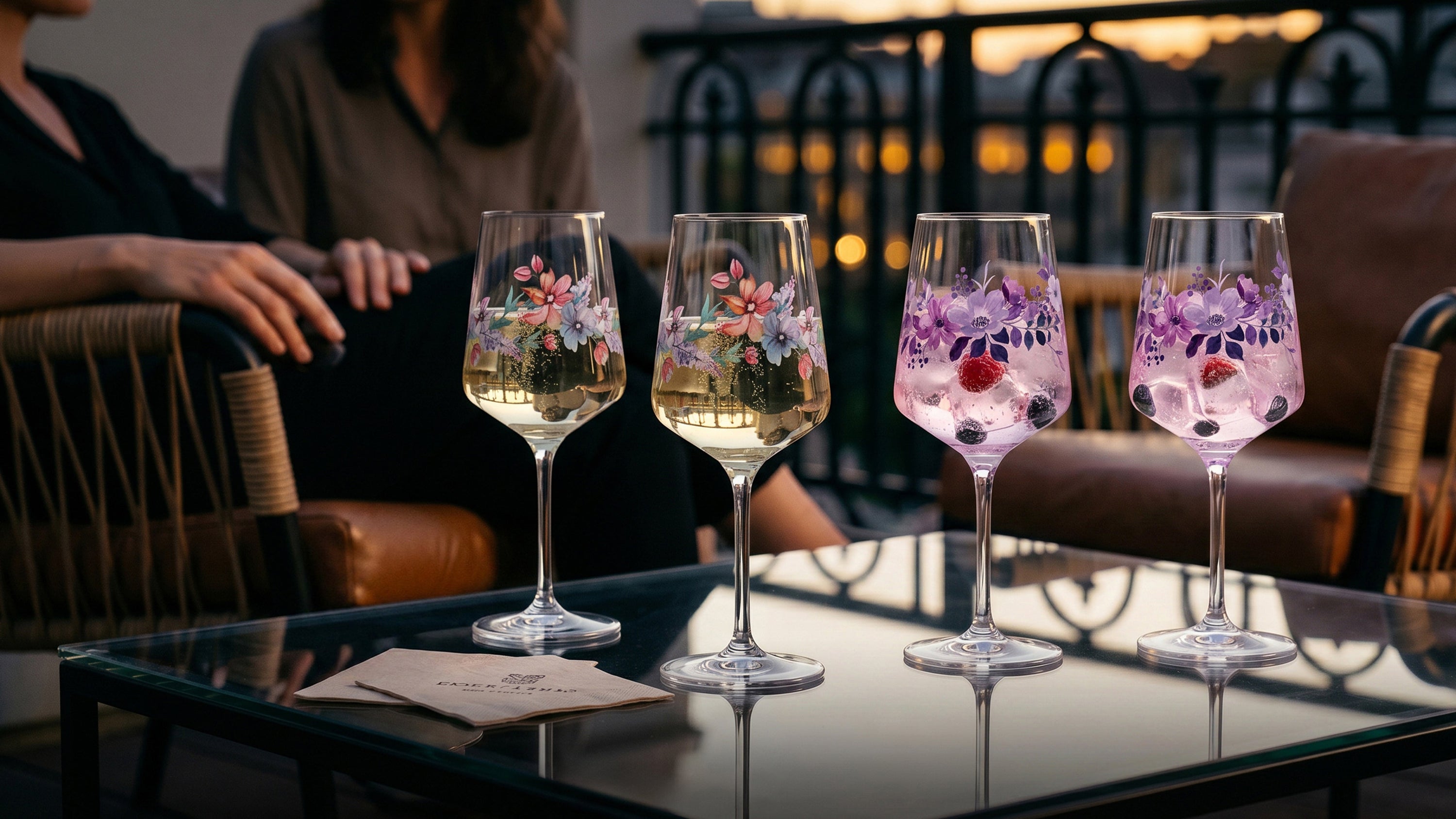 Vier ElbFuchs Aperitifgläser mit floralen Mustern auf einem Glastisch: Zwei Gläser mit bunten Sommerblumen und zwei Gläser der Kollektion Violet Veil mit lila Blumenmuster, gefüllt mit spritzigen Cocktails und Beeren. Im Hintergrund sitzen zwei Frauen auf einer gemütlichen Dachterrasse während eines stimmungsvollen Sonnenuntergangs zur Abenddämmerung.