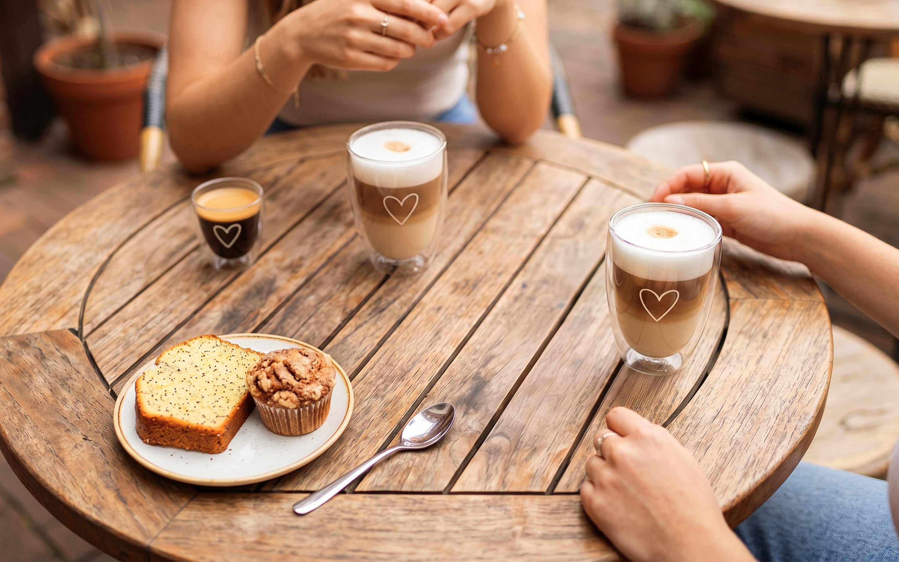 ElbFuchs PARIS doppelwandiges Kaffeeglas-Set aus Borosilikatglas mit feiner Herzgravur, bestehend aus einem 80ml Espresso-Glas und zwei 350ml Latte-Macchiato-Gläsern. Die Thermo-Gläser stehen zusammen mit einem Teller Kuchen auf einem runden, rustikalen Holz-Gartentisch. Zwei Personen genießen einen gemütlichen Kaffeemoment und ein bewusstes Kaffeeritual im Freien auf einer Terrasse.