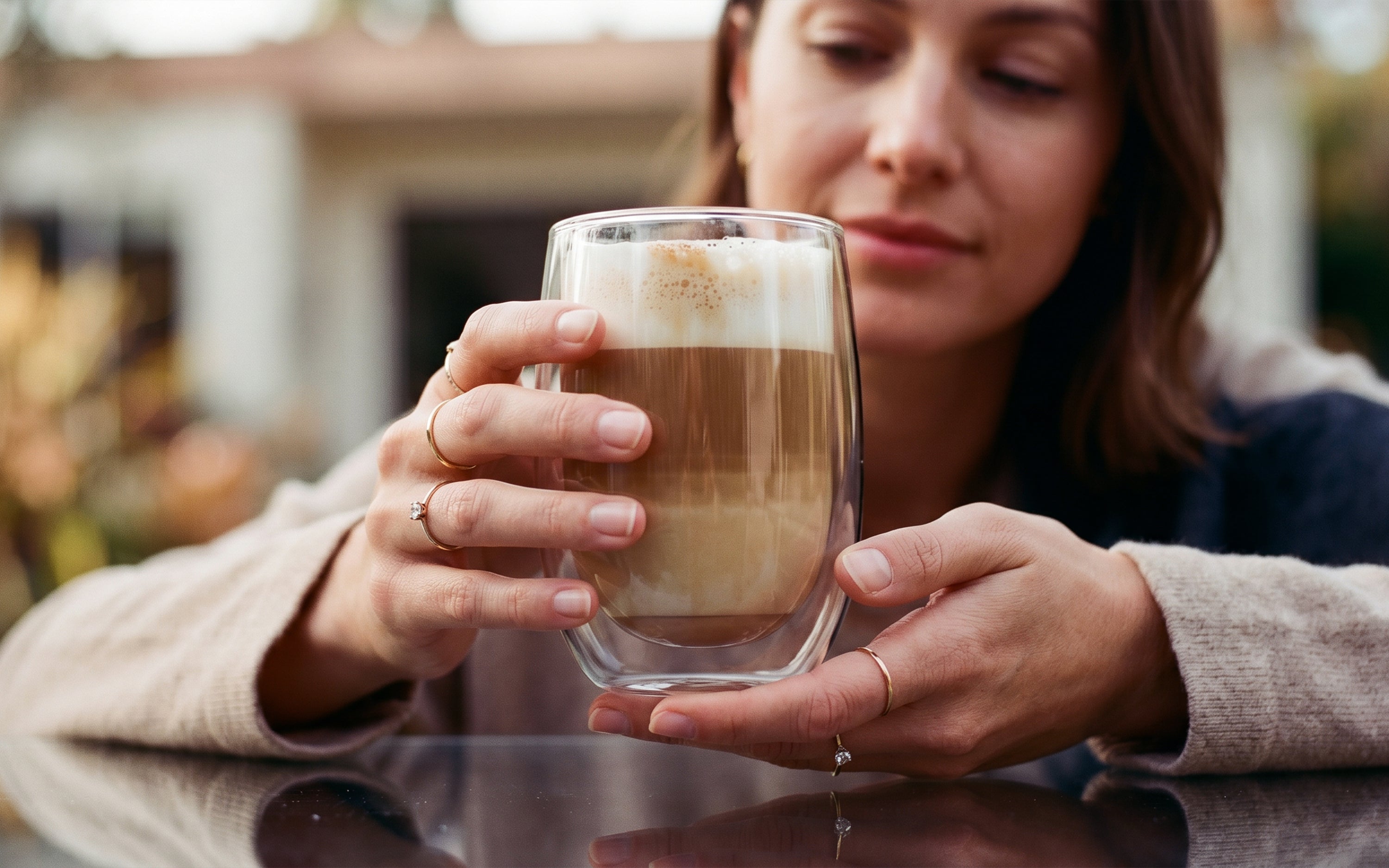 Eine Frau hält ein großes, doppelwandiges ElbFuchs Thermoglas aus der Kollektion 'MILAN' (350ml) behutsam mit beiden Händen. Das Kaffeeglas steht auf einer spiegelnden Tischoberfläche und ist mit einem ästhetisch geschichteten Latte Macchiato und cremigem Milchschaum befüllt. Die Frau trägt filigrane Ringe sowie einen gemütlichen hellen Strickpullover. Der sanft verschwommene Hintergrund lenkt den vollen Fokus auf den Kaffeegenuss und das zeitlose Design des Glases.