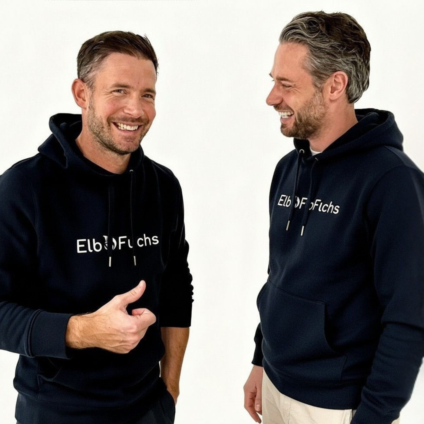 Die beiden ElbFuchs-Gründer Christoph und Kevin aus Hamburg lachen herzlich und sympathisch vor einem cleanen, weißen Studiohintergrund. Beide tragen passende dunkelblaue Hoodies mit dem weißen ElbFuchs-Markenlogo auf der Brust. Authentisches Team- und Gründerfoto.