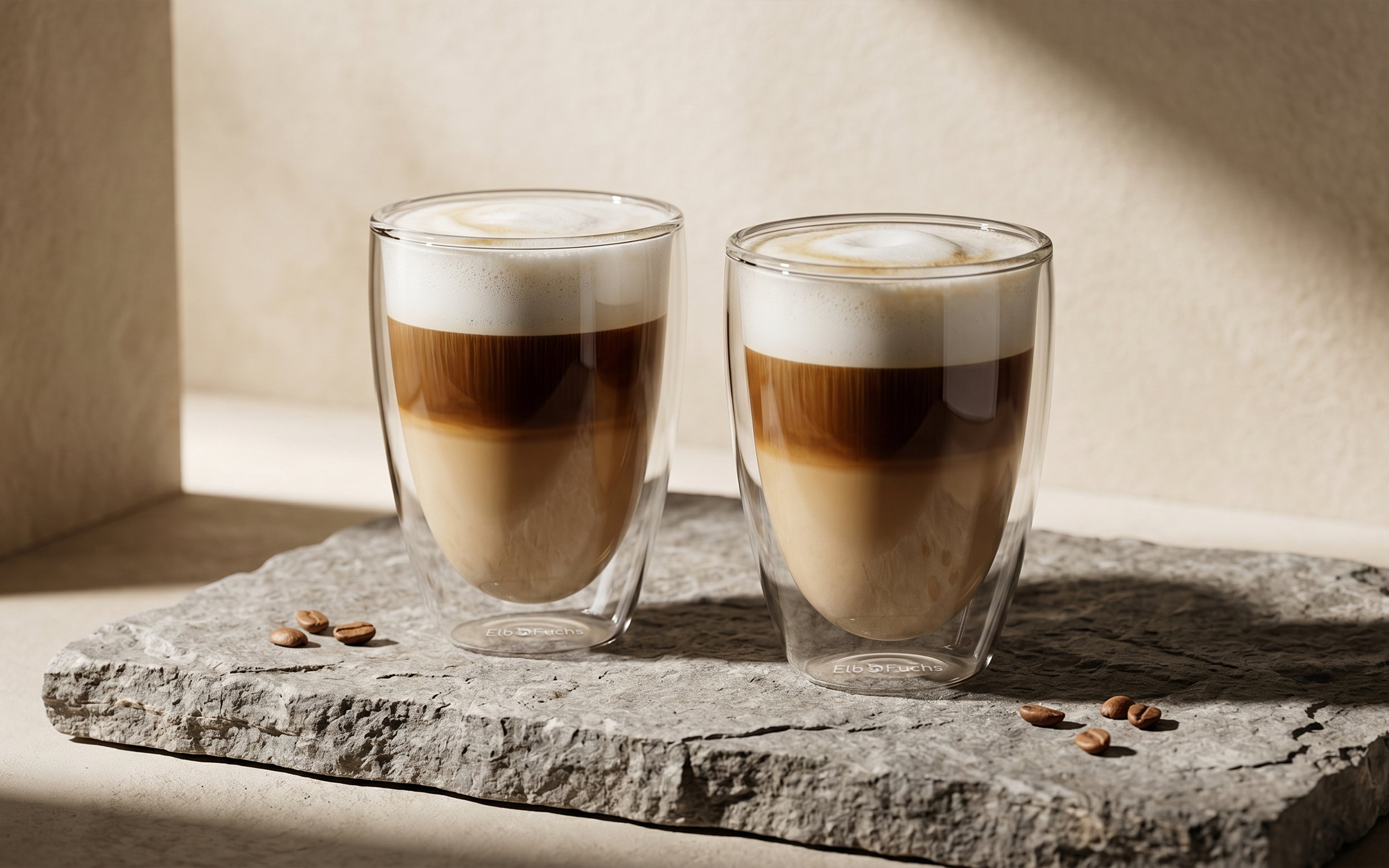 ElbFuchs VIENNA doppelwandiges Kaffeeglas-Set (350ml) aus Borosilikatglas, befüllt mit geschichtetem Latte Macchiato und cremigem Milchschaum. Die zwei Thermo-Kaffeegläser stehen auf einer rauen, grauen Natursteinplatte mit verstreuten Kaffeebohnen in einer hellen Umgebung mit elegantem Licht- und Schattenspiel.