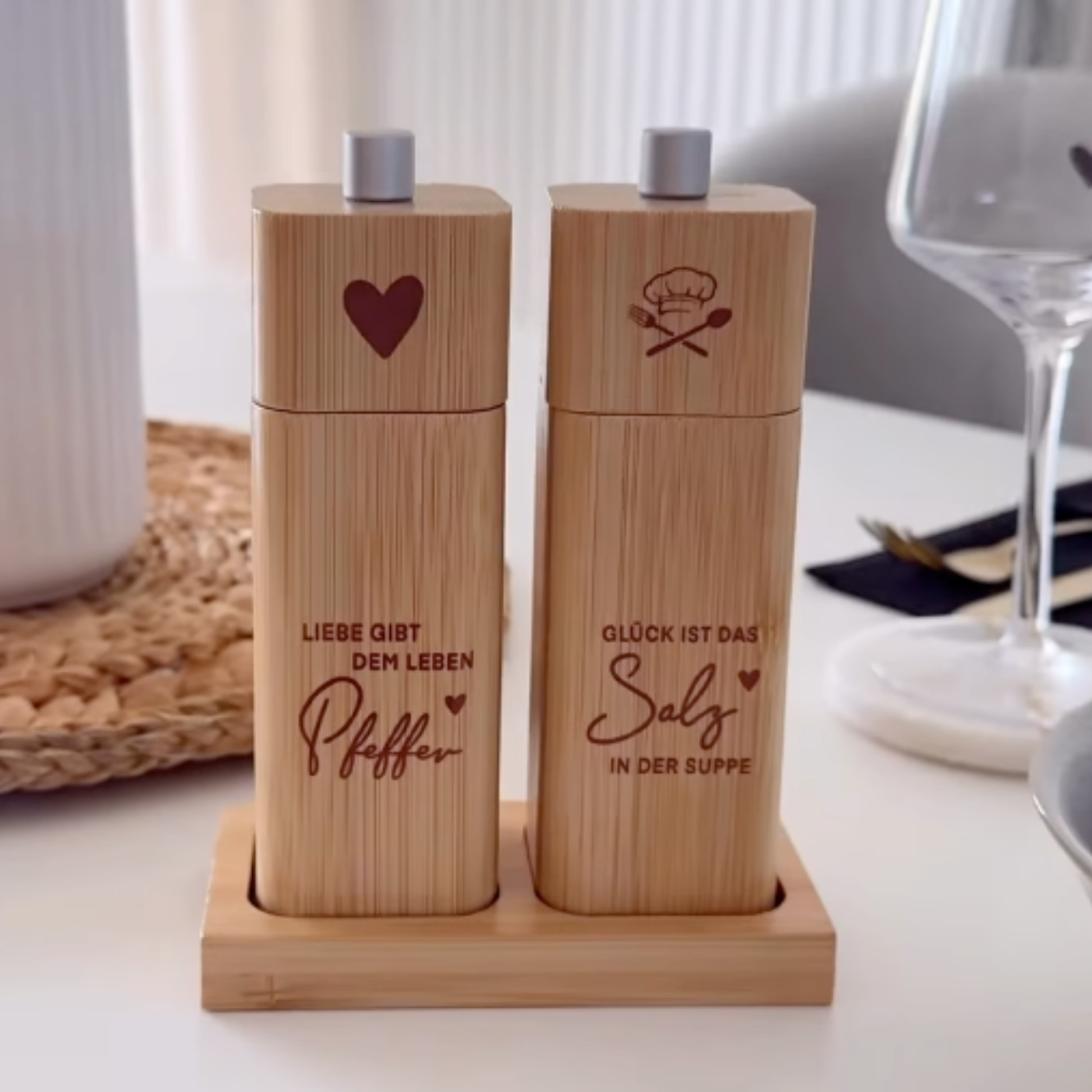 Ein ElbFuchs Salz- und Pfeffermühlen-Set aus hellem Bambusholz, das aufrecht in einer passenden Holzbasis auf einem weißen Tisch präsentiert wird. Die linke Mühle ist mit einem großen Herz-Symbol und dem gravierten Spruch 'LIEBE GIBT DEM LEBEN Pfeffer' versehen, während die rechte Mühle ein Kochmützen-Symbol sowie den Spruch 'GLÜCK IST DAS Salz IN DER SUPPE' trägt. Im weich gezeichneten, modernen Hintergrund sind ein Weinglas auf einem weißen Marmoruntersetzer.