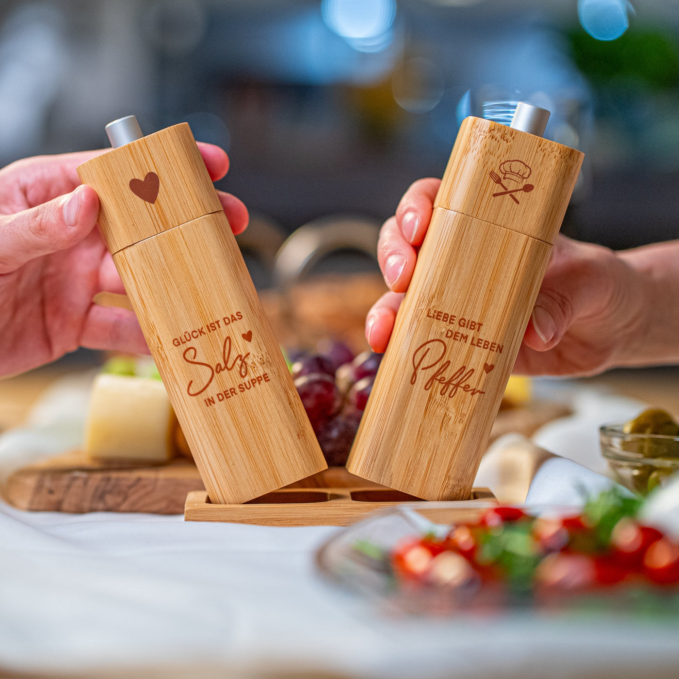 Ein ElbFuchs Salz- und Pfeffermühlen-Set aus hellem Bambusholz, das von zwei Händen über einer passenden Holzbasis gehalten wird. Die Salzmühle links ist mit einem Herz-Symbol und dem gravierten Spruch 'GLÜCK IST DAS Salz IN DER SUPPE' versehen. Die Pfeffermühle rechts zeigt eine Kochmütze mit gekreuztem Besteck und den Spruch 'LIEBE GIBT DEM LEBEN Pfeffer'. Im weich gezeichneten Hintergrund einer einladenden Tischszene sind Weintrauben, Käse und frischer Salat zu erkennen.