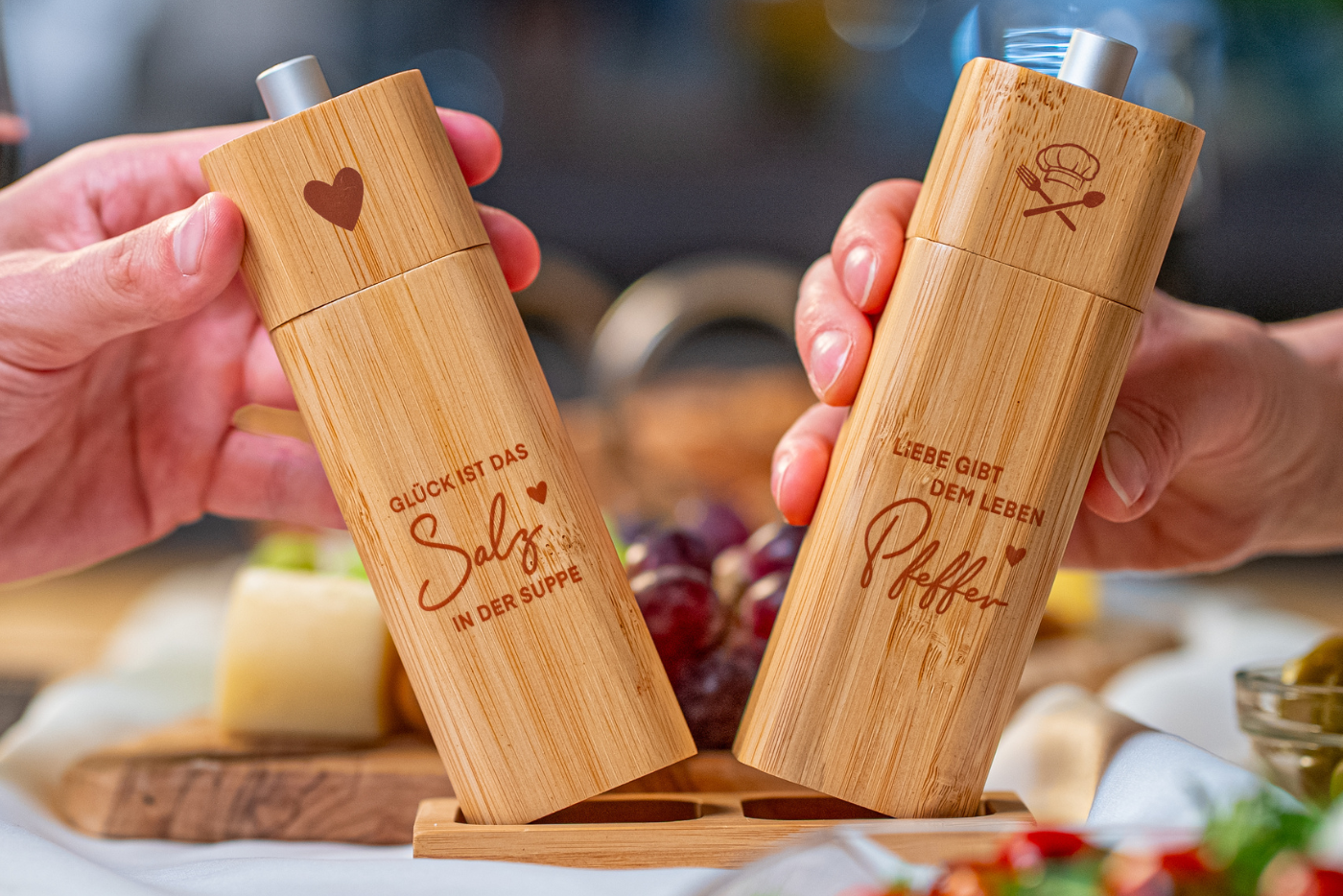 Ein ElbFuchs Salz- und Pfeffermühlen-Set aus hellem Bambusholz, das von zwei Händen über einer passenden Holzbasis gehalten wird. Die Salzmühle links ist mit einem Herz-Symbol und dem gravierten Spruch 'GLÜCK IST DAS Salz IN DER SUPPE' versehen. Die Pfeffermühle rechts zeigt eine Kochmütze mit gekreuztem Besteck und den Spruch 'LIEBE GIBT DEM LEBEN Pfeffer'. Im weich gezeichneten Hintergrund einer einladenden Tischszene sind Weintrauben, Käse und frischer Salat zu erkennen.