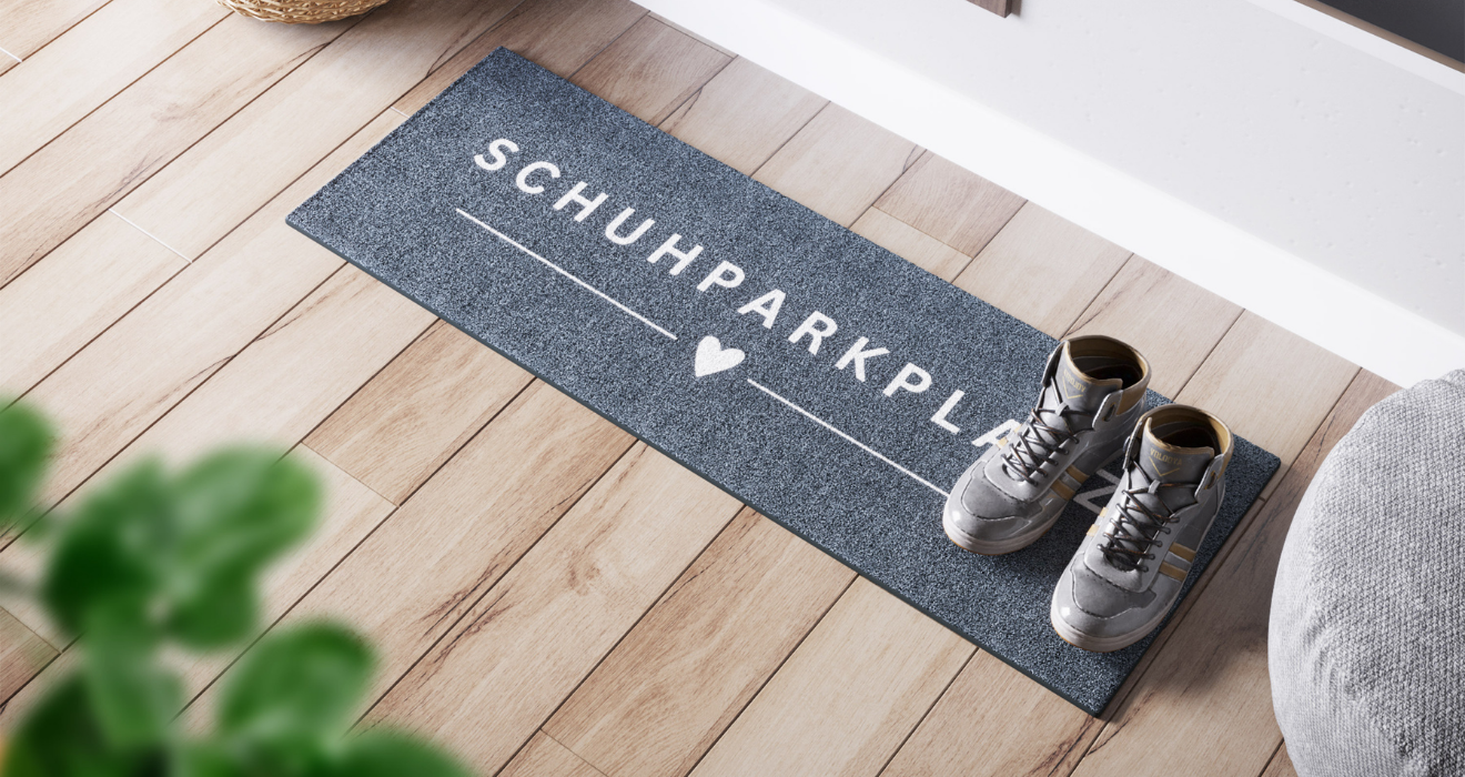 Schmale ElbFuchs Abtropfmatte "SCHUHPARKPLATZ" mit Herzsymbol aus Hamburg. 35x100 cm Schuhteppich auf Holzboden neben grauen Sneakern. Robuste und dekorative Fussmatte für Norddeutschland.