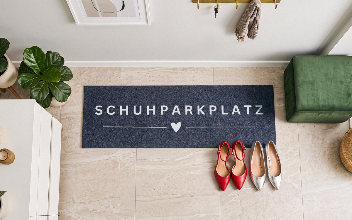 Anthrazite ElbFuchs Schuhmatte „SCHUHPARKPLATZ“ mit Herz-Motiv (100x35 cm) aus der Vogelperspektive im hellen Flur, platziert auf beigen Fliesen neben rotem und silbernem Schuhwerk, grünen Zimmerpflanzen und einer Samt-Sitzbank.