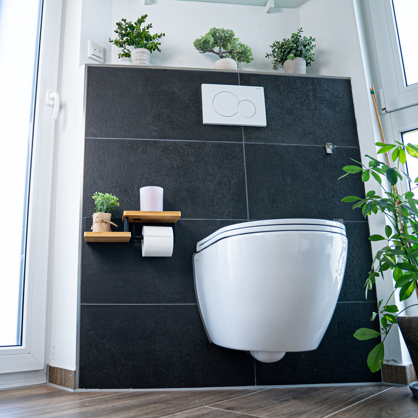 Ein modernes, helles Badezimmer-Ambiente mit einem eleganten, weißen Wand-WC vor einer kontrastreichen Wand aus großen, mattschwarzen Fliesen. Links neben der Toilette ist der ElbFuchs Toilettenpapierhalter 'Singapor' mit seinen zwei versetzten Bambus-Ablagen montiert, die mit einer kleinen Pflanze und einer Kerze dekoriert sind. Mehrere grüne Zimmerpflanzen auf dem oberen Fliesengesims und ein Boden in warmer Holzoptik runden das stilvolle Lifestyle-Produktfoto ab.