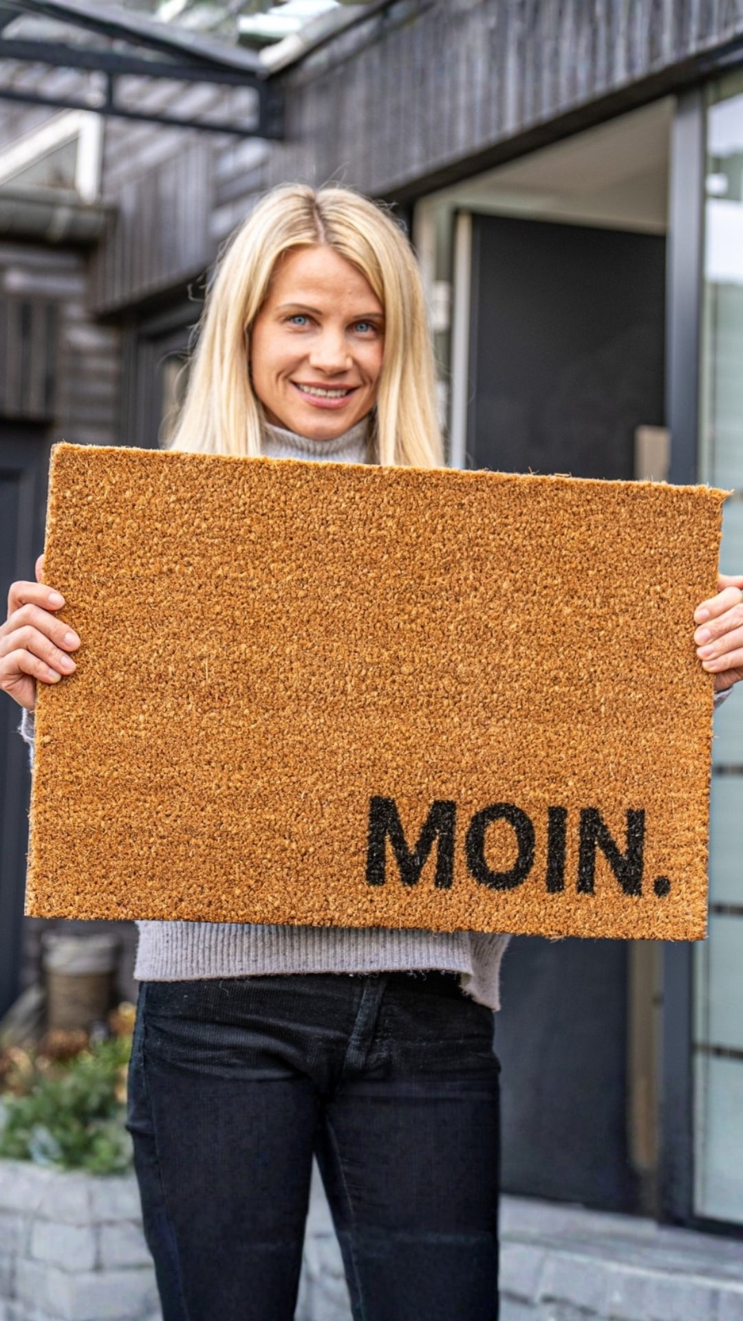 Frau hält die ElbFuchs Kokos-Fußmatte mit Schriftzug MOIN. (60x40 cm) vor einem modernen Hauseingang.