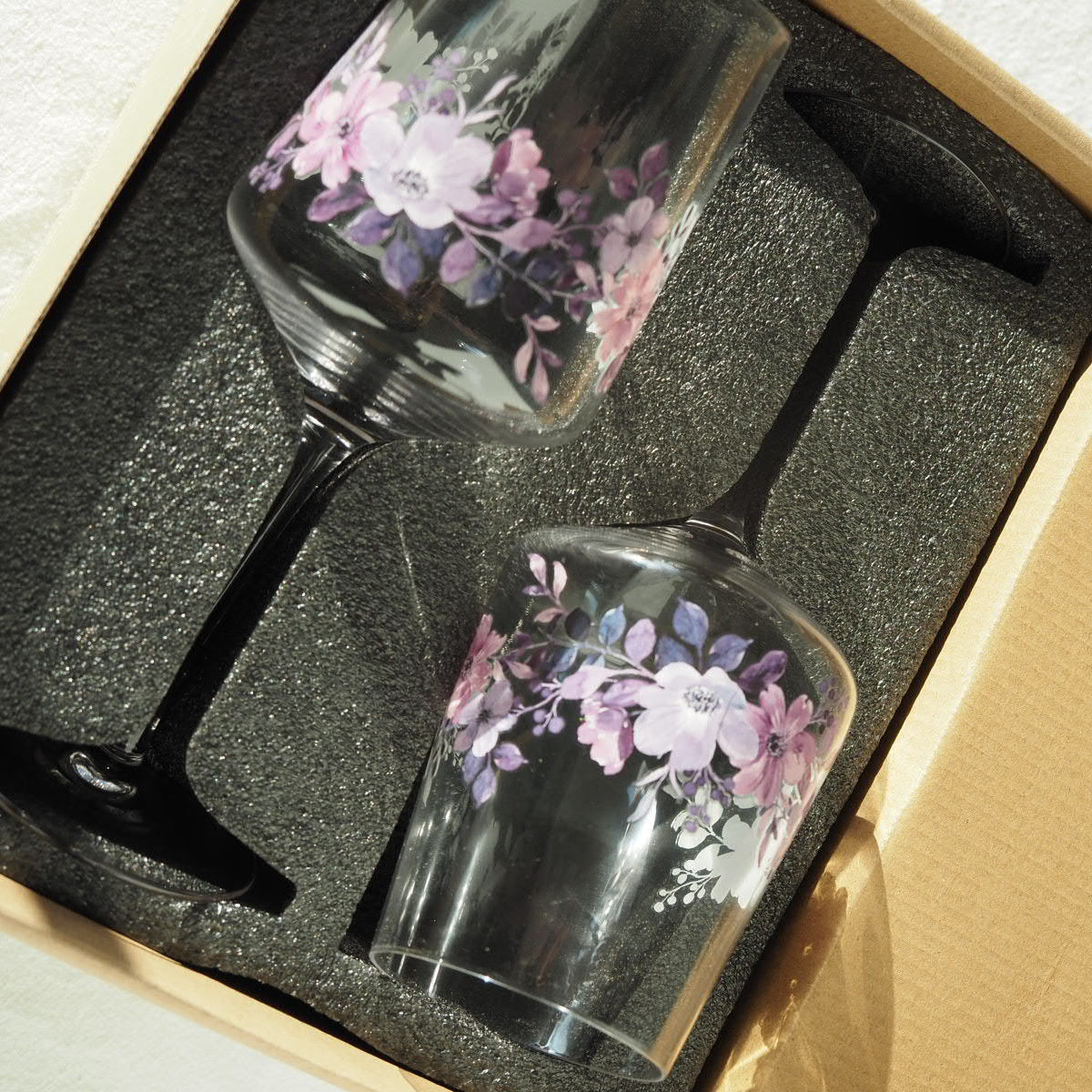 Zwei ElbFuchs Aperitifgläser (450ml) mit elegantem, violett-lilafarbenem Blumenmuster, eingebettet in eine passgenaue, bruchsichere Schaumpolsterung aus schwarzem Schaumstoff in einem braunen Karton. Die Nahaufnahme im hellen Sonnenlicht verdeutlicht den sicheren Transportschutz und die hochwertige Verpackung des Gläser-Sets für den sicheren Versand.