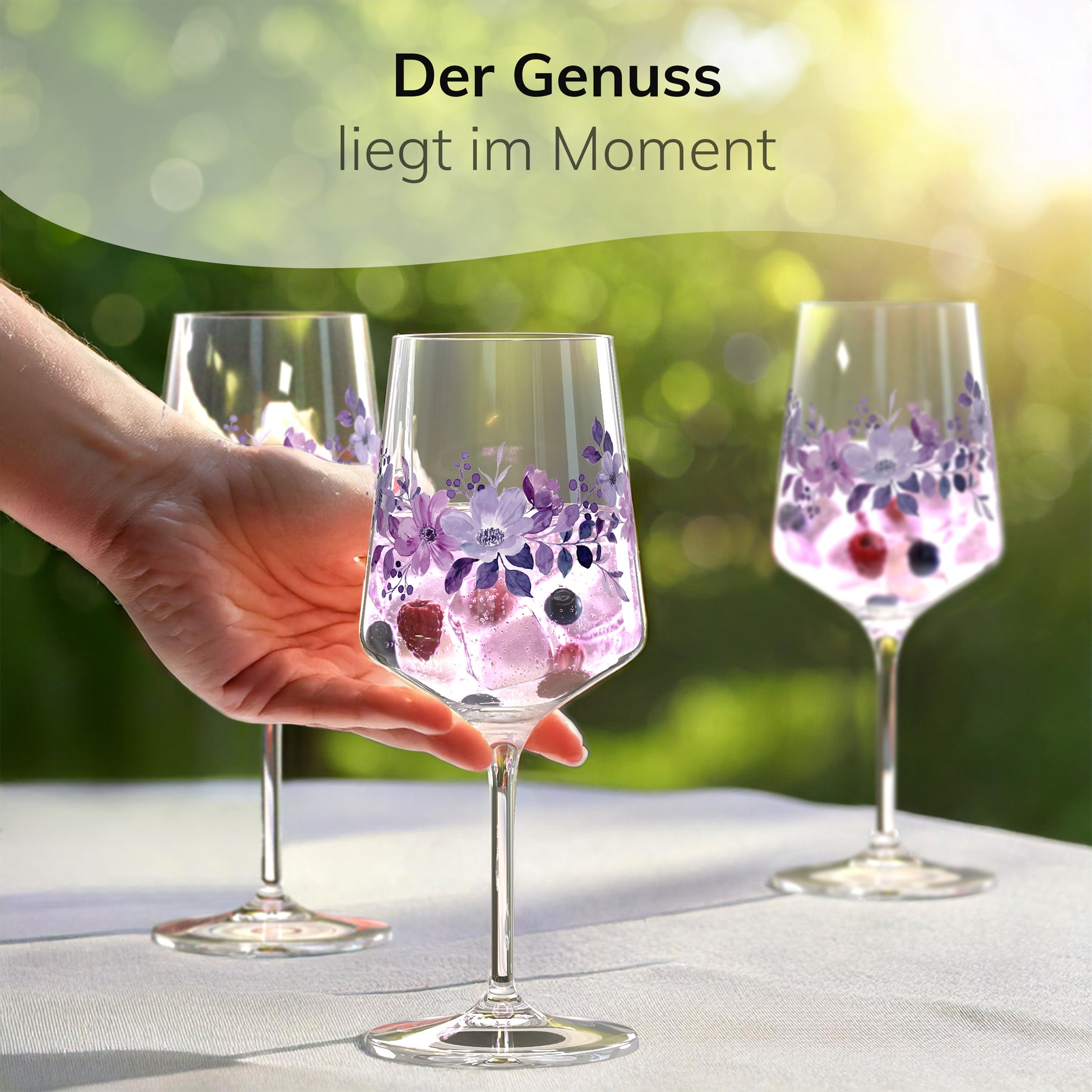 Eine Hand greift nach einem ElbFuchs Glas 'Violet Veil' mit violettem Blütenmotiv, gefüllt mit einem beerigen Getränk mit Himbeeren und Brombeeren, auf einem weiß gedeckten Gartentisch, im Hintergrund zwei weitere Gläser im Sonnenlicht.