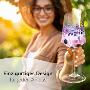 Eine lächelnde Frau mit Sonnenhut hält im Garten ein ElbFuchs Glas 'Violet Veil' mit violettem Blütenmotiv und einem beerigen, hellrosa Getränk in die Kamera, dahinter verschwommenes Grün im warmen Licht.