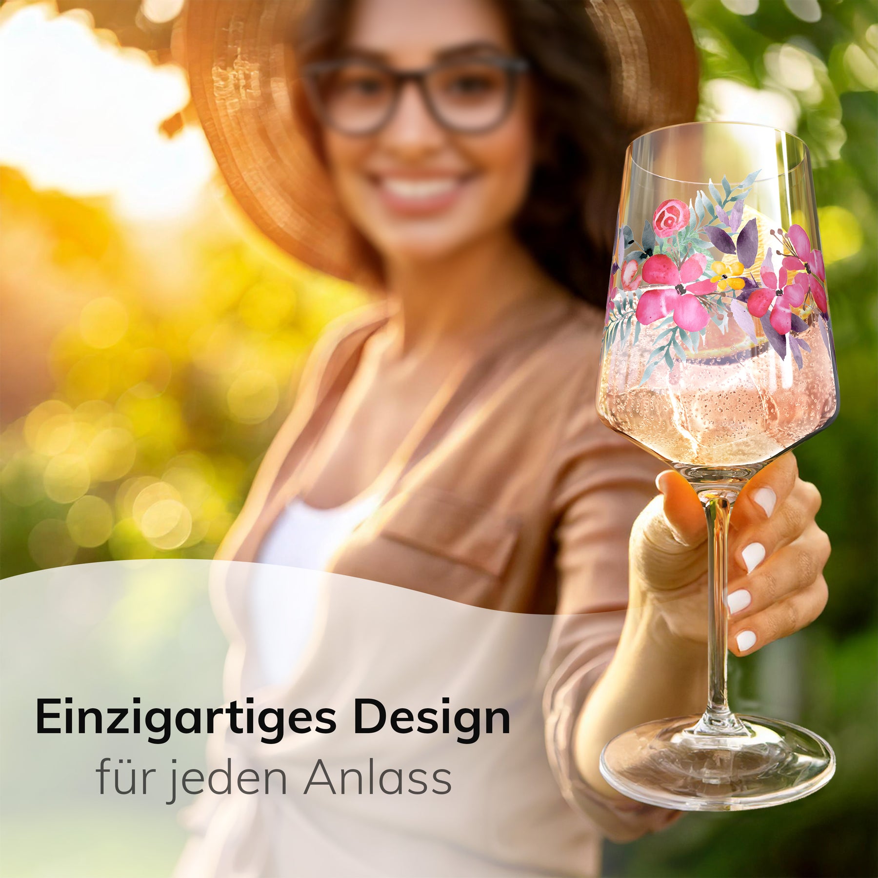 Eine lächelnde Frau mit Sonnenhut und Brille hält in sonnigem Garten ein ElbFuchs Aperitifglas 'Rosé Garden' mit zartem Blumenmotiv in die Kamera, gefüllt mit einem spritzigen, rosé-farbenen Aperitif vor grün verschwommenem Hintergrund.