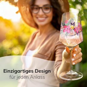 Eine lächelnde Frau mit Sonnenhut und Brille hält in sonnigem Garten ein ElbFuchs Aperitifglas 'Rosé Garden' mit zartem Blumenmotiv in die Kamera, gefüllt mit einem spritzigen, rosé-farbenen Aperitif vor grün verschwommenem Hintergrund.