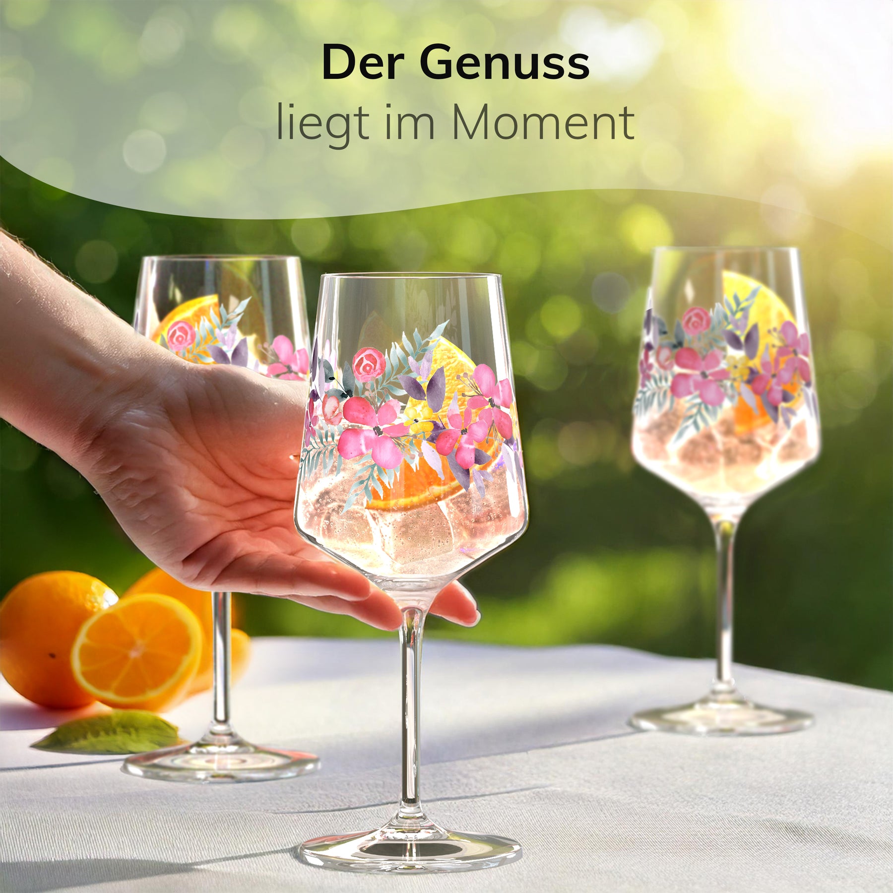 Drei ElbFuchs Aperitifgläser mit floralem Rosé-Garden-Motiv stehen mit einem Orangen-Spritz und Eiswürfeln gefüllt auf einer weißen Tischdecke im sonnigen Garten, während eine Hand nach einem der Gläser mit frischen Orangenscheiben daneben greift.