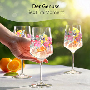 Drei ElbFuchs Aperitifgläser mit floralem Rosé-Garden-Motiv stehen mit einem Orangen-Spritz und Eiswürfeln gefüllt auf einer weißen Tischdecke im sonnigen Garten, während eine Hand nach einem der Gläser mit frischen Orangenscheiben daneben greift.