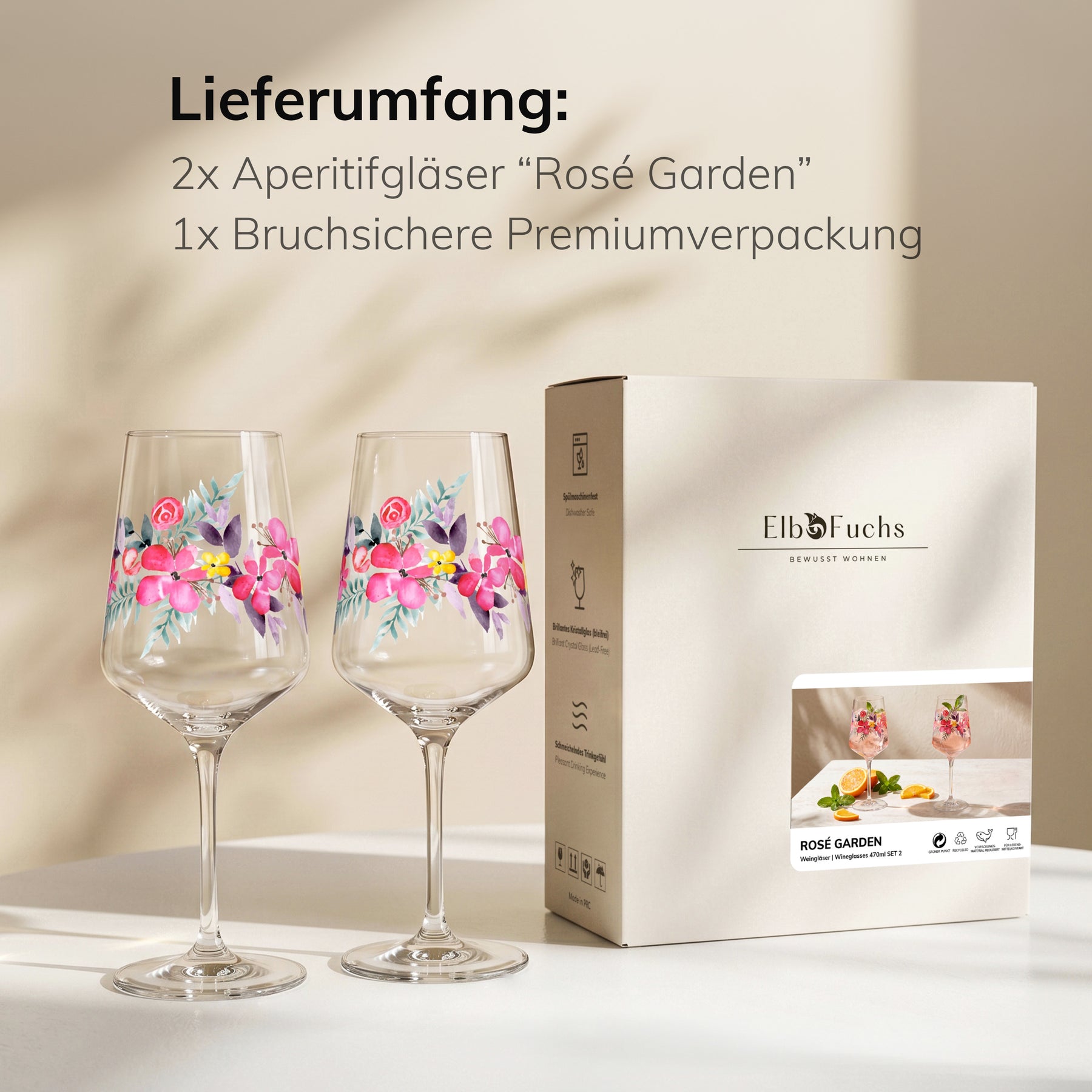 ElbFuchs Geschenkset 'Rosé Garden': zwei mit Rosenblüten bemalte Weingläser stehen neben der Verpackung mit Produktfoto auf einem hellen Tisch im warmen Streiflicht.