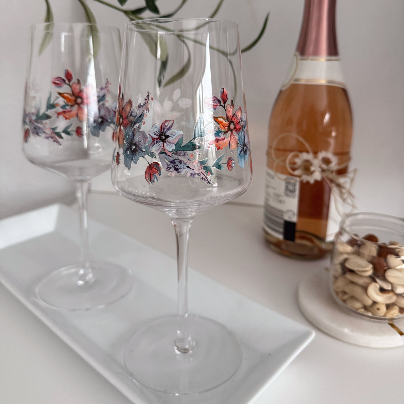 Zwei elegante ElbFuchs Aperitifgläser aus der Kollektion 'Spring Harmony' (470ml) mit einem detailreichen, farbenfrohen Blumenmuster in Korallenrot, Blau und Lila. Die langstieligen Gläser stehen auf einer länglichen, weißen Servierplatte. Im weich gezeichneten Hintergrund vervollständigen eine Flasche Rosé-Sekt und ein kleines Knabberglas mit Nüssen auf einem runden Marmoruntersetzer die gemütliche Genussszene.