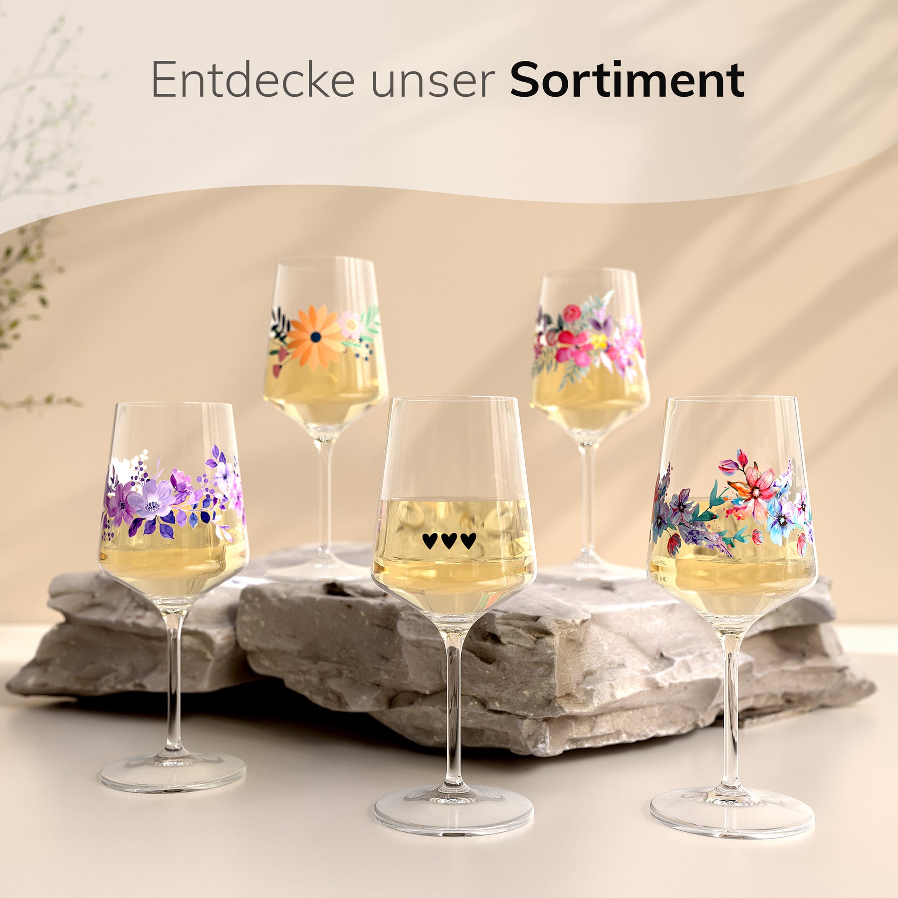 Fünf verschiedene ElbFuchs Weingläser mit unterschiedlichen floralen Motiven und einer Herzchen-Gravur, gefüllt mit Weißwein und arrangiert auf einem hellen Steinuntergrund unter dem Schriftzug 'Entdecke unser Sortiment'.