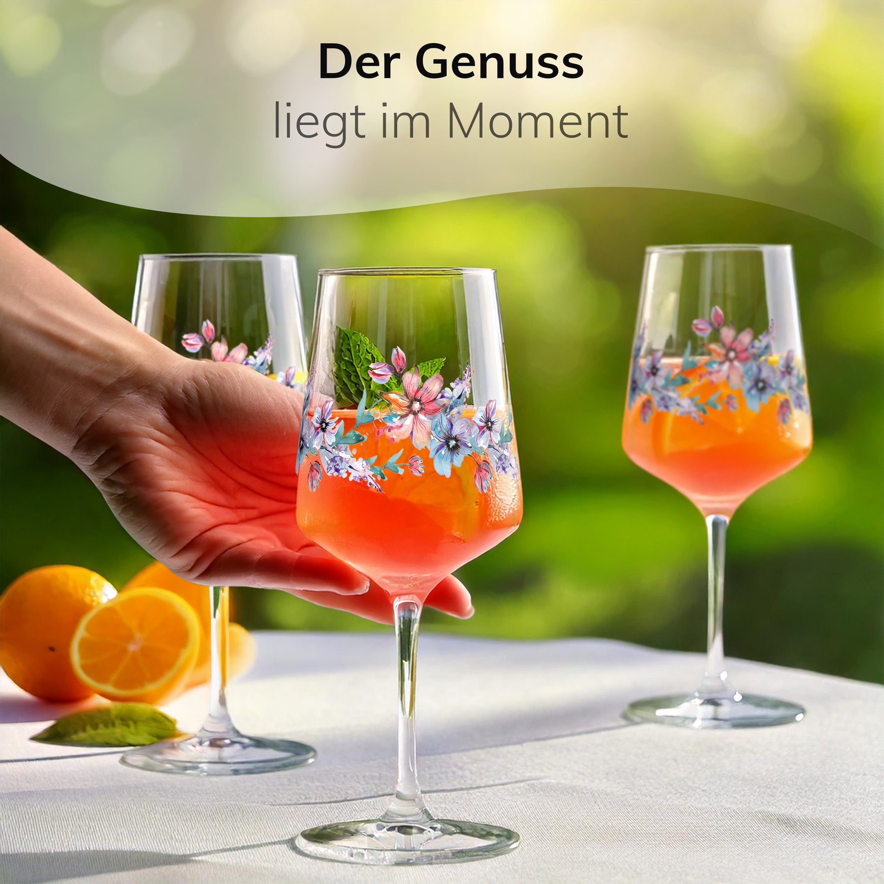 Eine Hand reicht ein mit Minze und Orange dekoriertes ElbFuchs Glas 'Spring Harmony' über einen gedeckten Gartentisch mit frischen Orangen, im Hintergrund zwei weitere blumenverzierte Gläser mit orangefarbenem Aperitif in sonniger Umgebung.