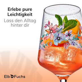 Nahaufnahme eines beschlagenen ElbFuchs Glases 'Spring Harmony' mit orangefarbenem Getränk, Eiswürfeln und frischer Minze, verziert mit zartem Blütenmuster, vor cremefarbenem Verlaufshintergrund.