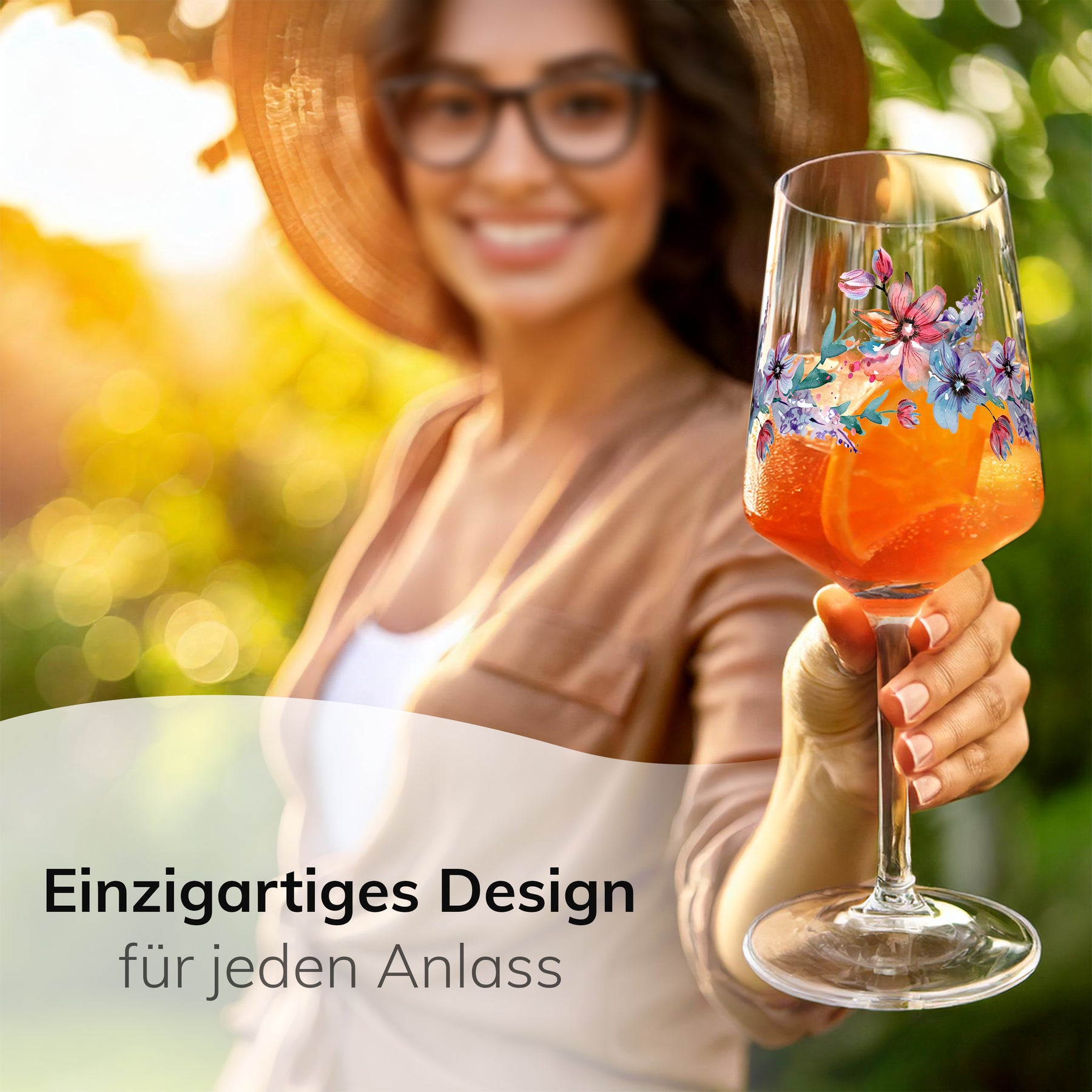 Eine lächelnde Frau mit Strohhut hält im sonnigen Garten ein ElbFuchs Glas 'Spring Harmony' mit orangefarbenem Aperitif und bunter Blütenverzierung in die Kamera, umgeben von grünem Blattwerk im Gegenlicht.