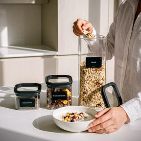 Frau füllt Müsli aus einem ElbFuchs BLACK KONTOR Vorratsglas in eine Schale, daneben kleine 400-ml-Gläser mit Chiasamen und Trockenfrüchten.