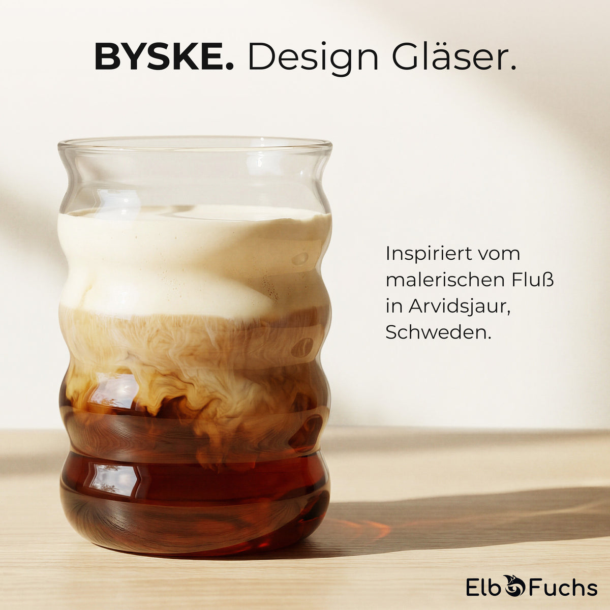 Ein ElbFuchs BYSKE Designglas mit geschichtetem Iced Coffee auf einem Holztisch im Sonnenlicht, der Text verweist auf die Inspiration durch den malerischen Fluss in Arvidsjaur, Schweden.