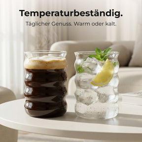 Zwei ElbFuchs BYSKE Gläser auf einem Couchtisch im Wohnzimmer, eines gefüllt mit dunklem Iced Coffee, das andere mit Infused Water aus Eiswürfeln, Minze und Zitrone, dazu der Hinweis auf die Temperaturbeständigkeit der Gläser.