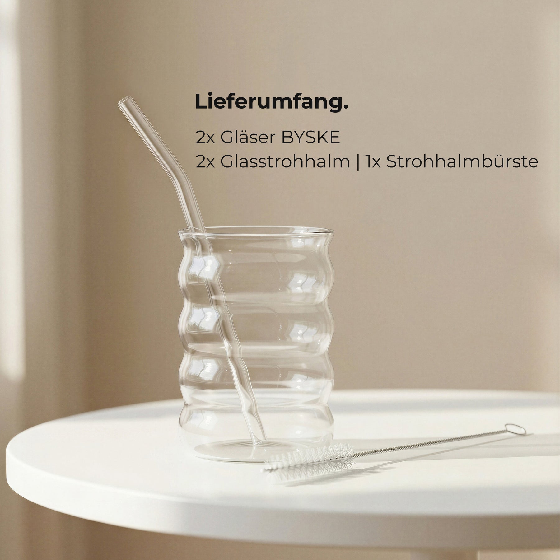 Ein leeres ElbFuchs BYSKE Glas mit wellenförmiger Riffelung, Glasstrohhalm und Reinigungsbürste auf einem weißen Tisch, daneben der Lieferumfang: 2 Gläser, 2 Glasstrohhalme und eine Strohhalmbürste.