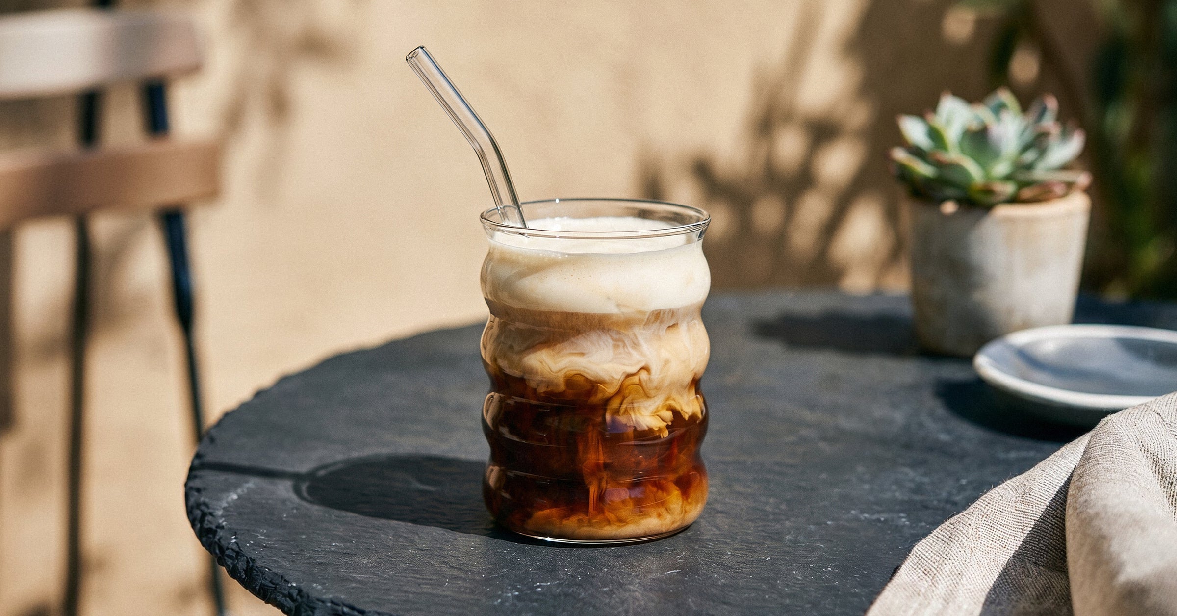 Ein Latte Macchiato serviert in einem Bubble-Glas mit Strohhalm von ElbFuchs, angerichtet auf einem schwarzen Schiefertisch mit leichter Sommerdeko an einem sonnigen Tag.