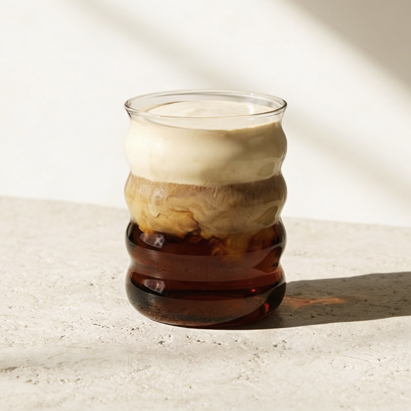 Ein ElbFuchs Bubbleglas (BYSKE) 400ml, gefüllt mit einem geschichteten Eiskaffee- oder Latte-Getränk, steht auf einem hellen Steintisch unter natürlichem Sonnenlicht und wirft einen harmonisch beruhigenden Schatten auf den minimalistischen Tisch.
