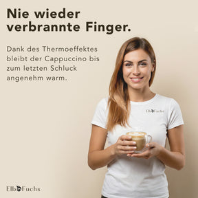 Eine lächelnde Frau im weißen ElbFuchs T-Shirt hält ein doppelwandiges VIENNA Kaffeeglas mit Henkel, gefüllt mit schaumigem Cappuccino, in beiden Händen. Der Bildtext verspricht dank Thermoeffekt angenehm warmen Kaffee bis zum letzten Schluck.