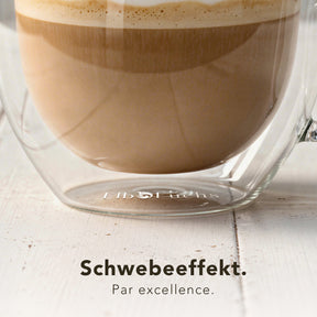 Nahaufnahme eines doppelwandigen ElbFuchs VIENNA Kaffeeglases, gefüllt mit Milchkaffee, auf einem hellen Holztisch. Die doppelte Wandung erzeugt den schwebenden Effekt, für den die Gläser bekannt sind.