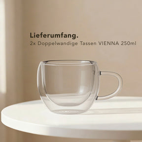 Ein leeres doppelwandiges ElbFuchs VIENNA Kaffeeglas mit Henkel aus Borosilikatglas steht auf einem runden weißen Tisch, dahinter der Hinweis auf den Lieferumfang von zwei Tassen à 250 ml.