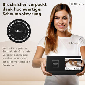 Eine lächelnde Frau im weißen ElbFuchs T-Shirt hält eine schwarze VIENNA Geschenkbox mit Zufriedenheitssiegel in den Händen, deren stoßfeste Schaumpolsterung die doppelwandigen Kaffeegläser sicher vor Bruch beim Versand schützt.