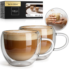 Zwei doppelwandige VIENNA Kaffeegläser mit Henkel, gefüllt mit cremigem Milchkaffee, stehen nebeneinander vor der ElbFuchs Verpackung im Set 2x250ml, ergänzt durch ein rundes Foto zweier Hände, die ein Glas umschließen.