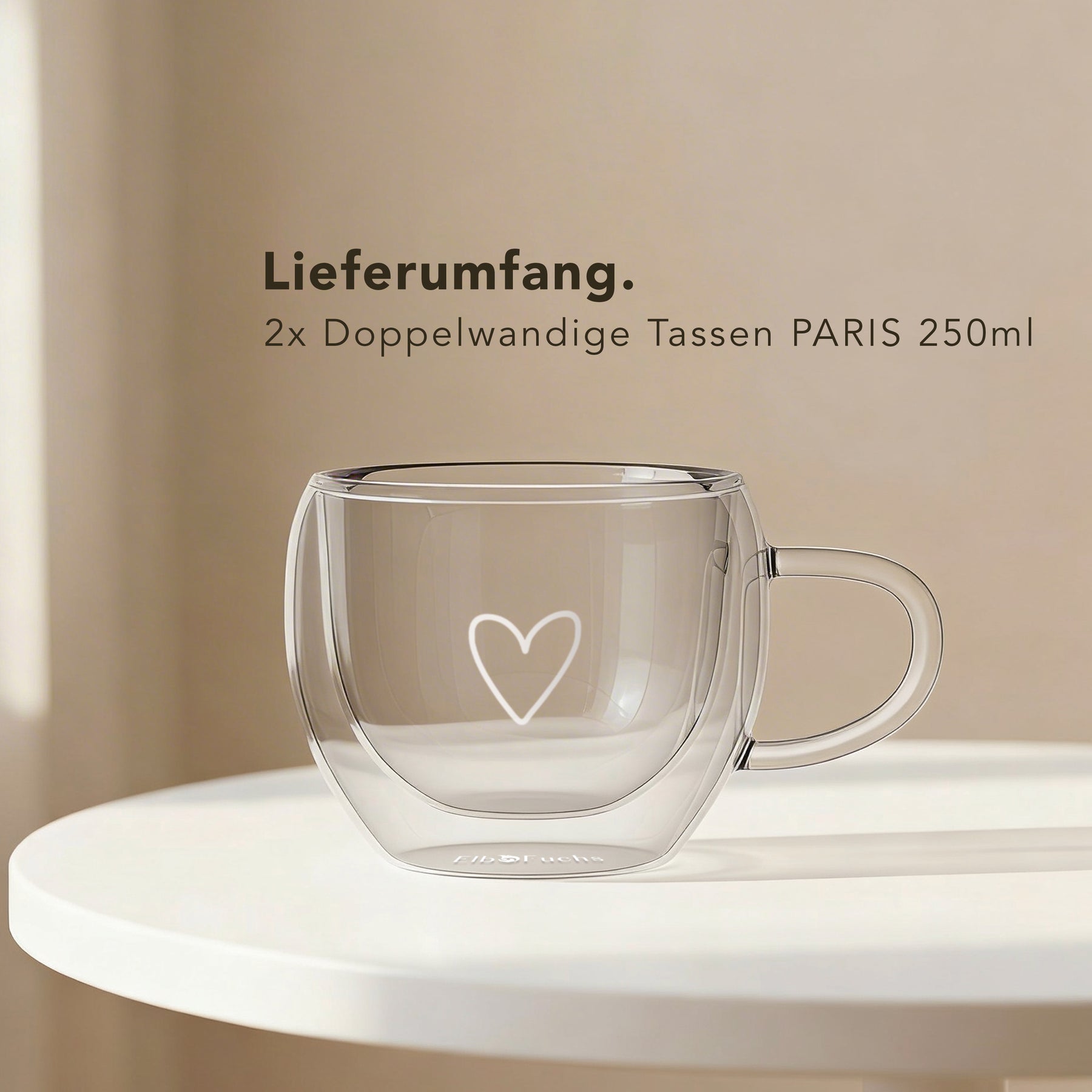 Ein leeres doppelwandiges ElbFuchs PARIS Kaffeeglas mit Henkel und Herzgravur aus Borosilikatglas steht auf einem runden weißen Tisch, dahinter der Hinweis auf den Lieferumfang von zwei Tassen à 250 ml.