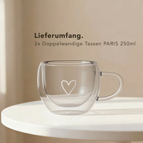 Ein leeres doppelwandiges ElbFuchs PARIS Kaffeeglas mit Henkel und Herzgravur aus Borosilikatglas steht auf einem runden weißen Tisch, dahinter der Hinweis auf den Lieferumfang von zwei Tassen à 250 ml.