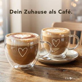 Zwei doppelwandige ElbFuchs PARIS Kaffeegläser mit Henkel und Herzgravur stehen gefüllt mit Cappuccino und kunstvoller Latte-Art auf einem Holztisch, eines davon auf einer weißen Untertasse mit Löffel, im warmen Licht einer gemütlichen Café-Atmosphäre.