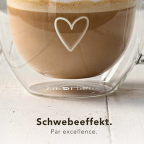 Nahaufnahme eines doppelwandigen ElbFuchs PARIS Kaffeeglases mit herzförmiger Gravur, durch das der doppelte Glasboden den charakteristischen Schwebeeffekt zeigt. Das Glas steht auf einem hellen Holztisch und ist mit Cappuccino und feinem Milchschaum gefüllt.