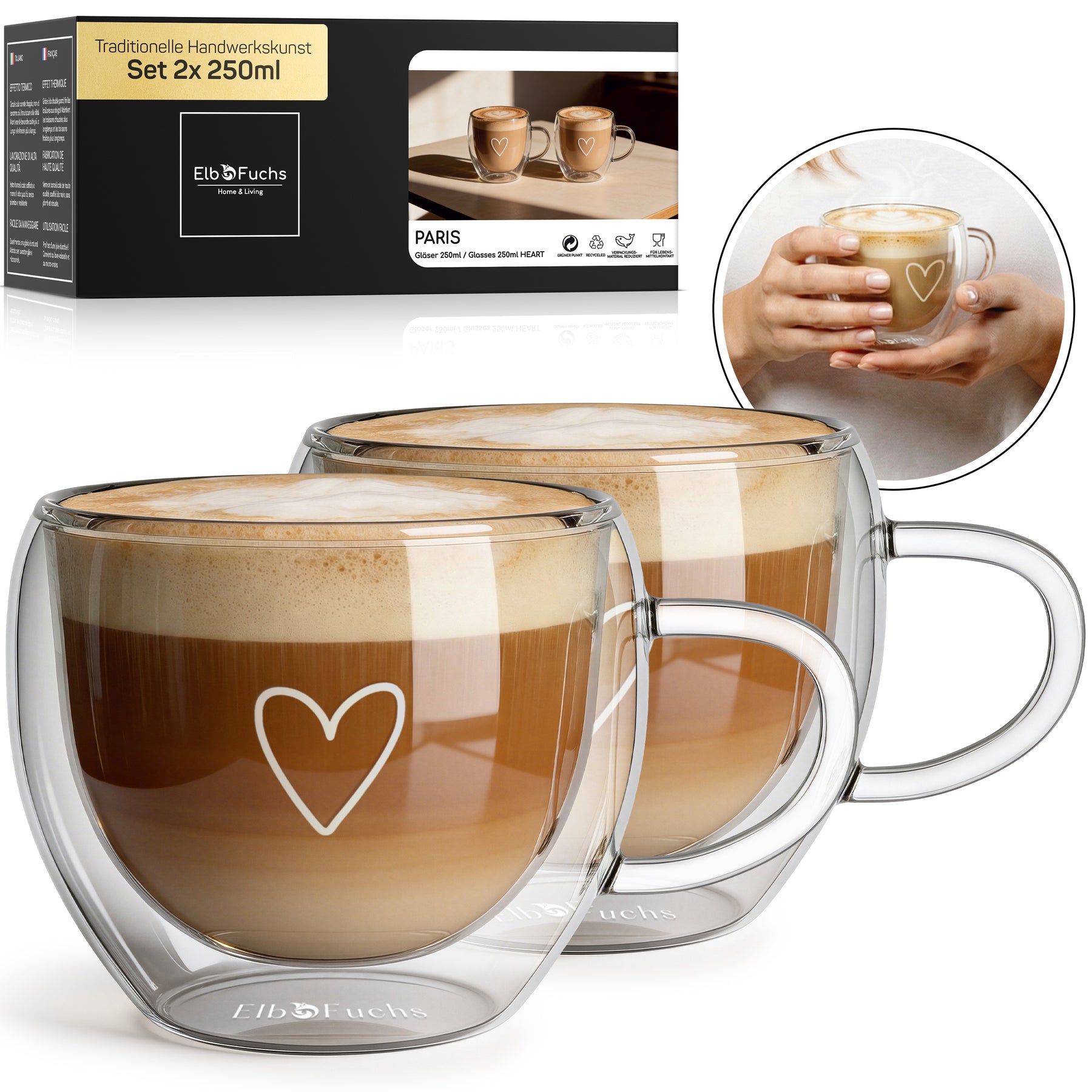 Zwei ElbFuchs PARIS Cappuccino-Tassen mit doppelwandigem Glas, Henkel und feiner Herzgravur, gefüllt mit cremigem Milchkaffee im Set 2x 250ml, mit Verpackung und einer Nahaufnahme haltender Hände im Hintergrund.