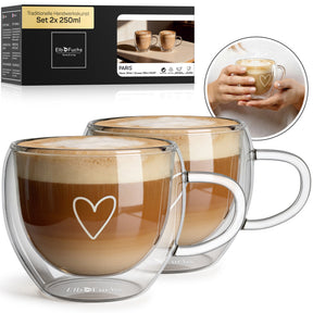 Zwei ElbFuchs PARIS Cappuccino-Tassen mit doppelwandigem Glas, Henkel und feiner Herzgravur, gefüllt mit cremigem Milchkaffee im Set 2x 250ml, mit Verpackung und einer Nahaufnahme haltender Hände im Hintergrund.