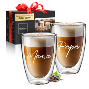 Vier doppelwandige ElbFuchs Gläser mit Latte Macchiato und feinem Milchschaum stehen auf einem Holztisch zwischen Kaffeebohnen, Keksen und einem Stück Kuchen, das mittlere Glas ziert die goldene Gravur 'Moin' neben einem Glas mit Anker-Motiv vor dunklem, stimmungsvollem Hintergrund.