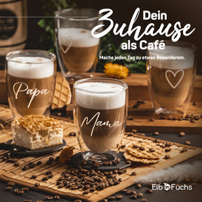 ElbFuchs Latte-macchiato-Gläser mit den Gravuren 'Papa' und 'Mama' auf einem rustikalen Holzbrett mit Kuchen, Keksen und Kaffeebohnen, dazu der Slogan 'Dein Zuhause als Café'.