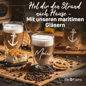 Maritime Tischszene mit ElbFuchs Thermogläsern, verziert mit Anker-Motiv und der Gravur 'Moin', gefüllt mit Latte macchiato und umgeben von Kaffeebohnen, Gebäck und einer Torte.