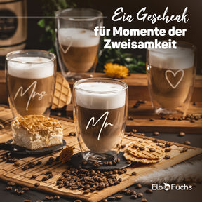 Drei doppelwandige ElbFuchs Latte-macchiato-Gläser mit den Gravuren 'Mrs', 'Mr' und einem Herzsymbol, dekoriert mit Kuchen, Keksen und Kaffeebohnen auf einer Bambusunterlage – ein Geschenk für Momente der Zweisamkeit.