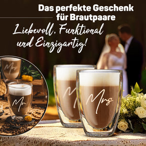 Zwei doppelwandige ElbFuchs Gläser mit den Gravuren 'Mr' und 'Mrs', gefüllt mit cremigem Latte macchiato, stehen vor einem unscharfen Brautpaar und einem Blumenstrauß als Hochzeitsgeschenk-Idee.