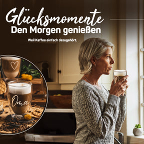 Eine ältere Frau in einem grauen Strickpullover trinkt genüsslich aus einem doppelwandigen ElbFuchs Glas mit der Gravur 'Oma' am Küchenfenster, im Bildausschnitt ist das Glas mit cremigem Latte macchiato zu sehen.