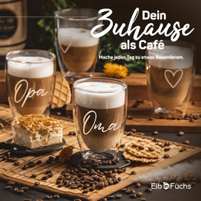 ElbFuchs Latte-macchiato-Gläser mit den Gravuren 'Oma' und 'Opa' auf einem Holzbrett mit Kuchen, Keksen und Kaffeebohnen, dazu der Slogan 'Dein Zuhause als Café'.